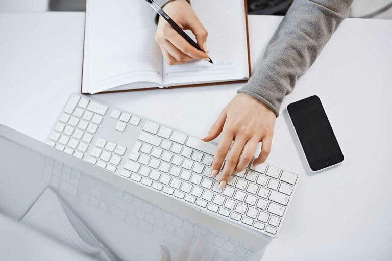 High angle shot of woman hands typing on keyboard. Focused female student working on her homework on computer, making notes, waiting for important call from boss and glancing at smartphone
